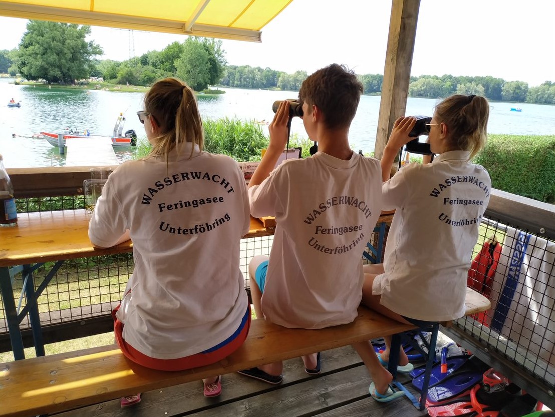 Drei Mitglieder mit Wasserwacht-T-Shirts gucken von einem Balkon auf einen See, zwei halten Ferngläser in der Hand.